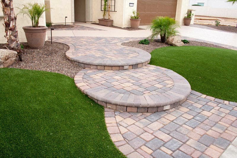 Curved paved walkway with grass and planters leading to a home entrance.