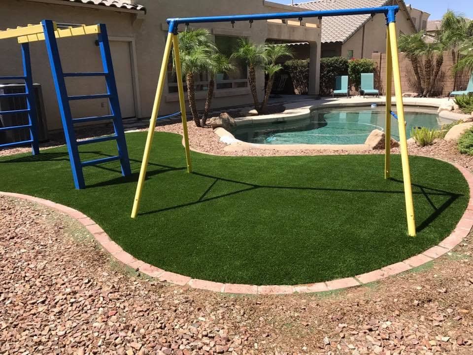 Backyard play area with yellow swingset, synthetic grass, and swimming pool surrounded by landscaping.