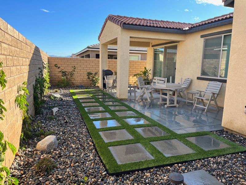 Modern backyard patio with stone pavers, artificial grass, and seating area under a shaded roof.