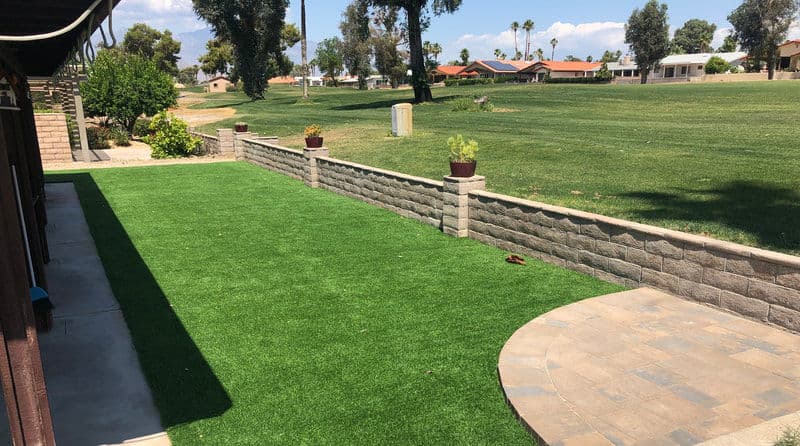 Lush green artificial turf lawn with a stone wall and landscaped plants near a golf course.