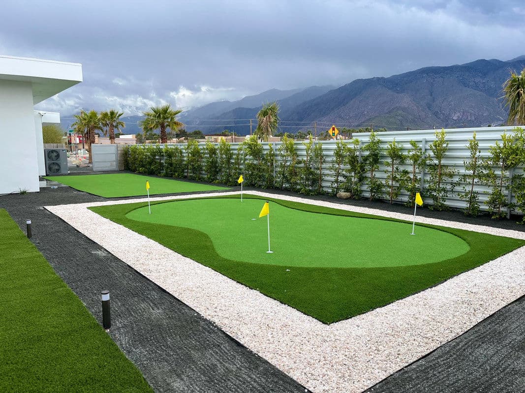 Artificial grass putting green with flags, surrounded by gravel and landscaping in mountainous setting.