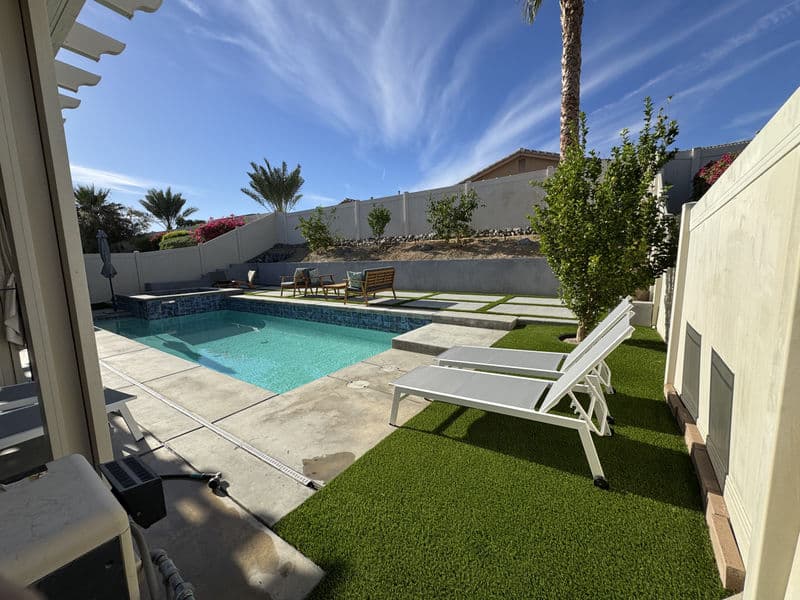 Modern backyard with a pool, lounge chairs, and lush greenery under a clear blue sky.