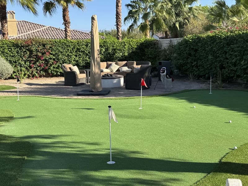 Artificial grass putting green with flags, patio furniture, and palm trees in a luxury backyard.