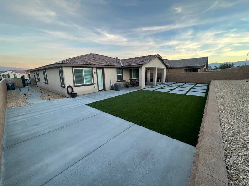 Modern backyard with artificial turf, concrete patio, and house under a scenic sky.